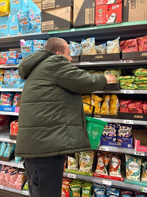 SEN Adult (Autism and Learning Diffculties) out shopping for his groceries, holding the basket and choosing the products himself while Staff stand aside for further support.